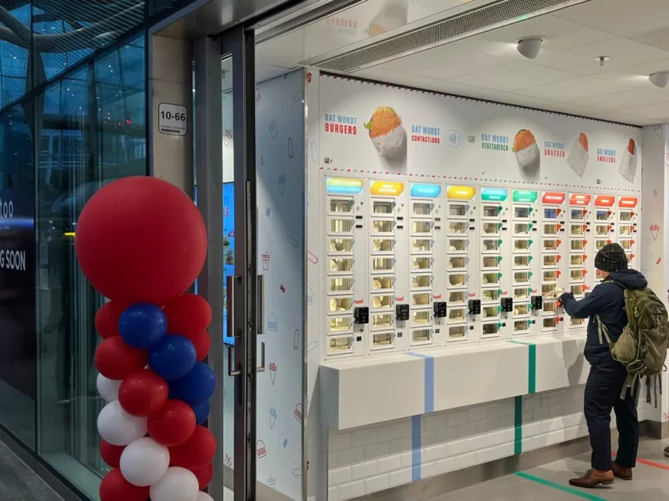 Een man met een rugzak neemt een snack uit de witte ADM-snackmuur bij Smullers